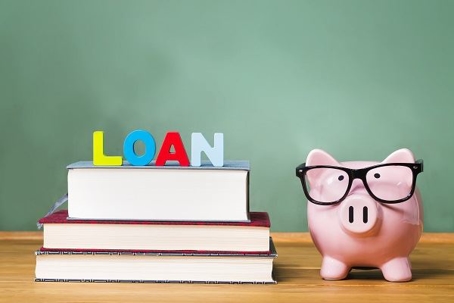 Stack of books on a desk with colored blocks that spell out "LOAN" next to a piggy bank with glasses