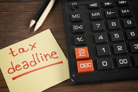 A yellow post it reads "tax deadline" in red marker next to a black calculator and some pencils